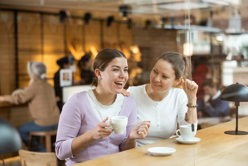 Two cheerful women chatting and drinking coffee at the bar counter of cafe