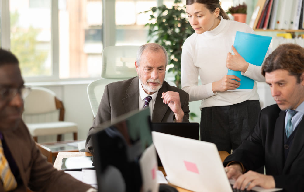 Woman with documents is standing next to senior male colleague with laptop