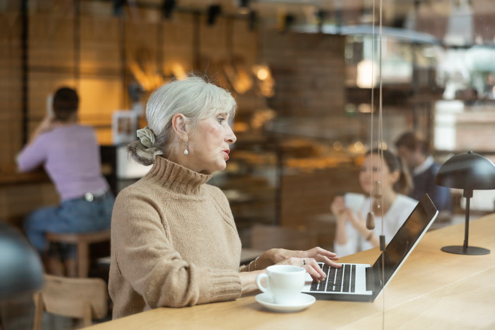 Positive European old woman drinking and enjoying coffee while working on laptop in cafe
