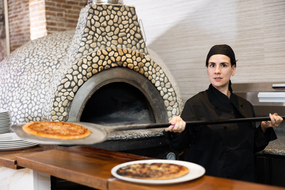 Latino female chef in black uniform holding big shovel with pizza in kitchen