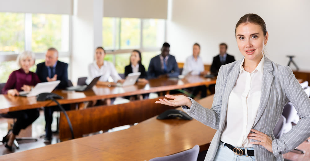 Female office employee standing at workplace in meeting room, making inviting gesture