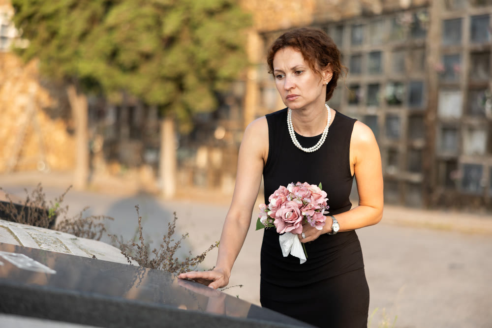 Sad woman in black dress with bouquet of flowers and handkerchief in hands, stands in cemetery