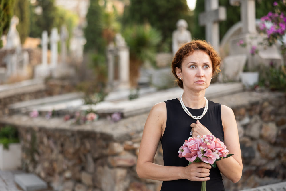 Woman in mourning black dress standing in cemetery with flowers