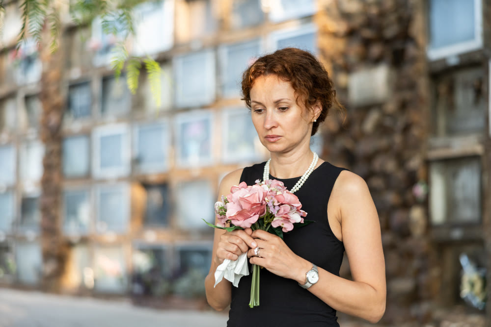 Sad woman in black dress with bouquet of flowers and handkerchief in hands, stands in cemetery