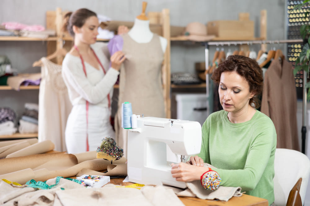 Female dressmaker sewing at machine while colleague working with mannequin