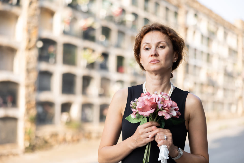 Sorrowful woman in black standing in cemetery with flowers