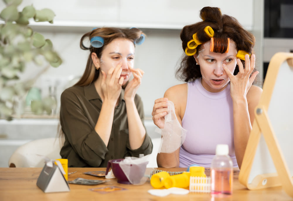 Female friends applying sheet masks to faces by vanity mirror
