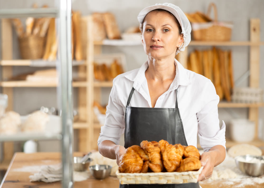 Female baker in white uniform stands behind counter in small private bakery offering hot croissants