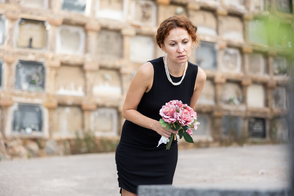 Upset woman in black dress, who lost loved one, brings flowers to grave