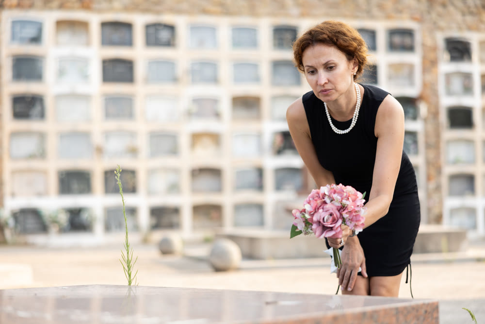 Sad widow in black lays bouquet of flowers on her dead husband grave
