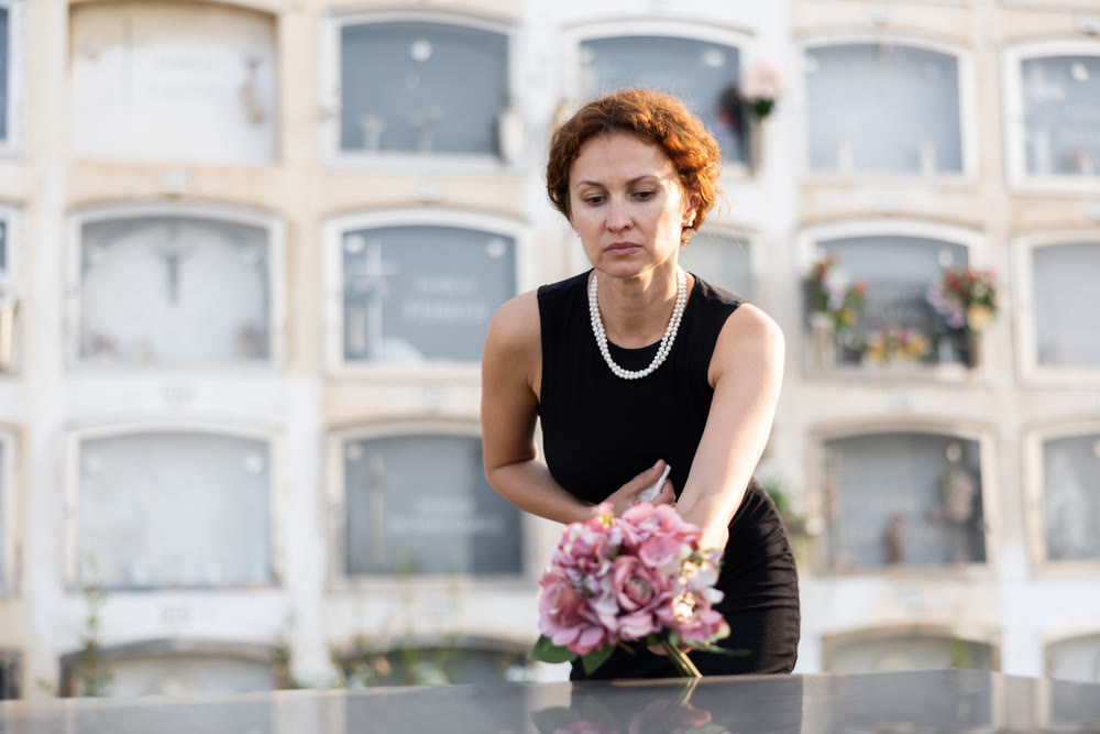 Sad widow in black lays bouquet of flowers on her dead husband grave