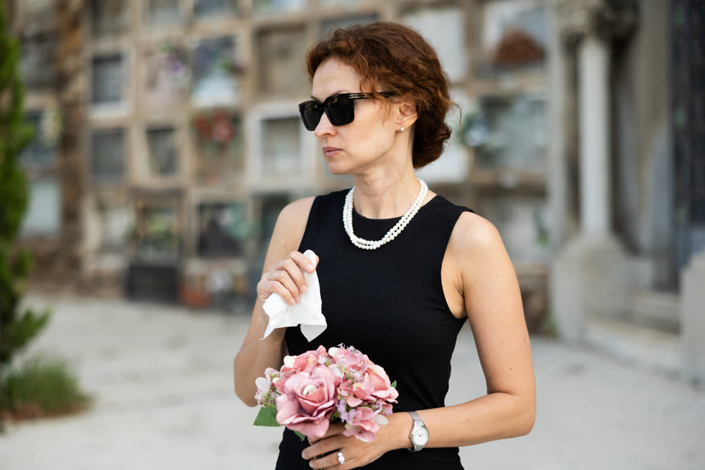 Sad woman in black dress with bouquet of flowers and handkerchief in hands, stands in cemetery