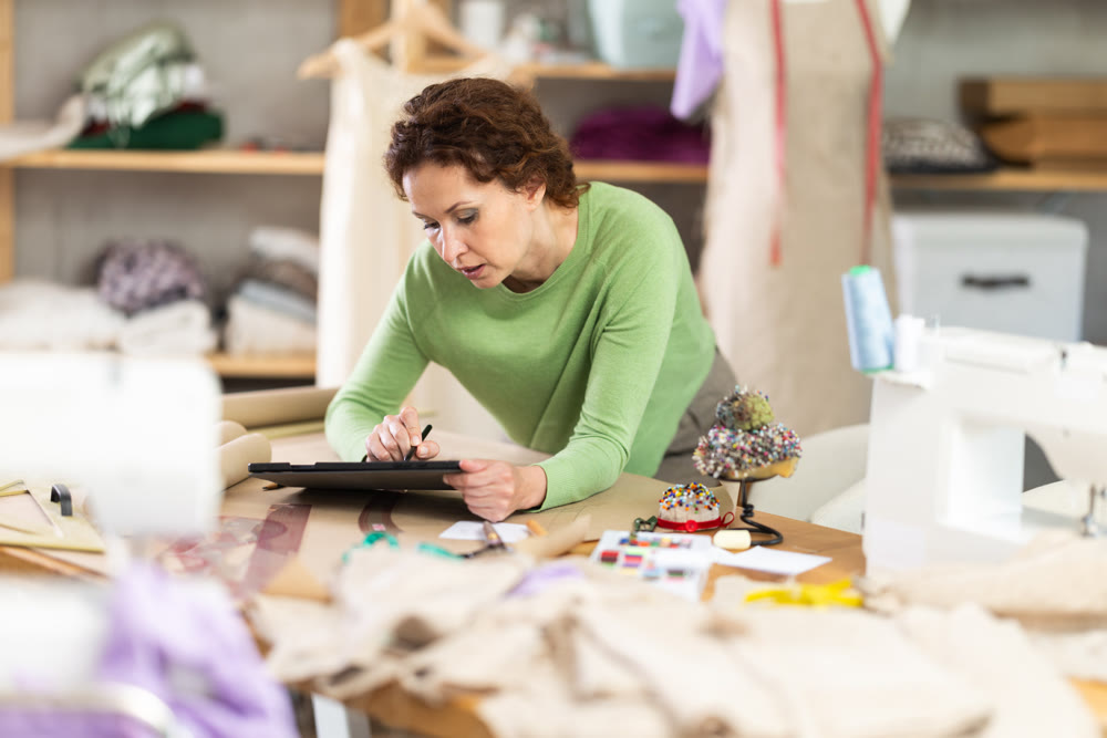 Female dressmaker draw outline of future dress on paper while looking at screen of an electronic tablet