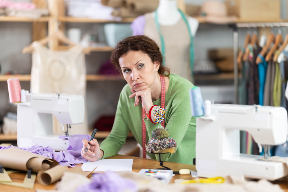 Thoughtful female tailor designs new clothes by drawing on white paper in workshop