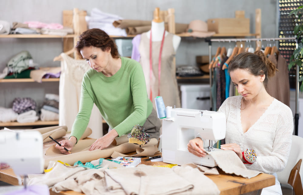 Female tailor sews on a sewing machine while another tailor draws outline of a future dress on paper