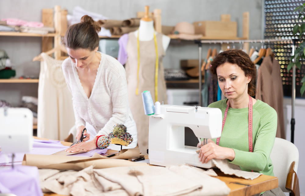 Female tailor sews on a sewing machine while another tailor cuts out patterns for dress with scissors