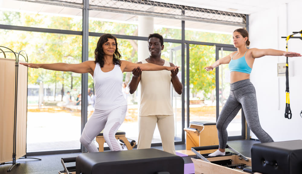 Attentive African American male trainer helping two positive women performing set of pilates
