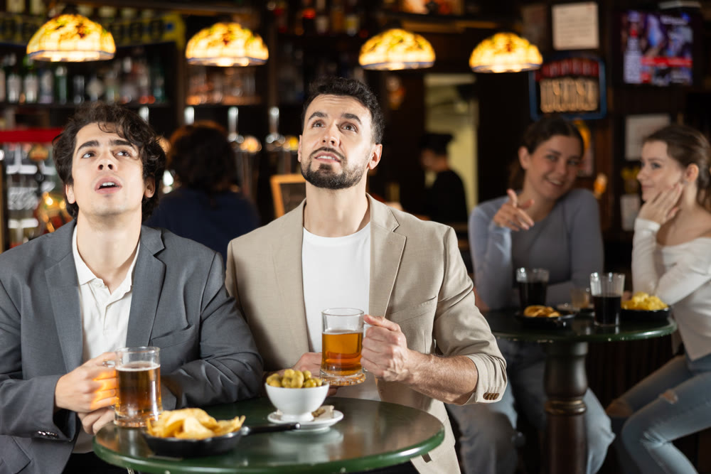 Two cheerful young men friends talking, watching match on TV and drinking beer at table in pub