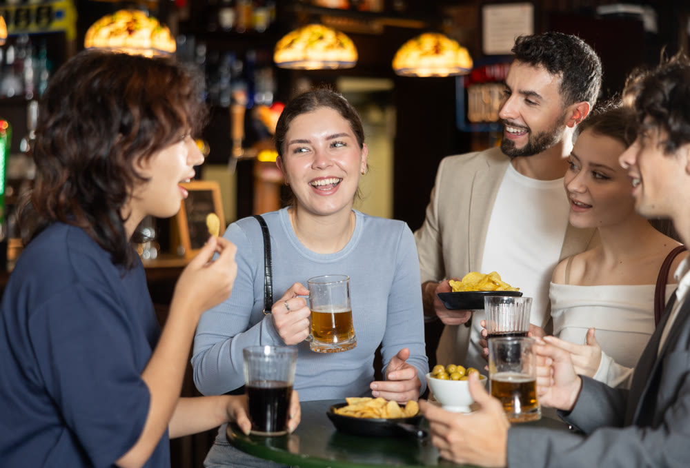 Group of positive young friends talking cheerfully and drinking beer at table in bar