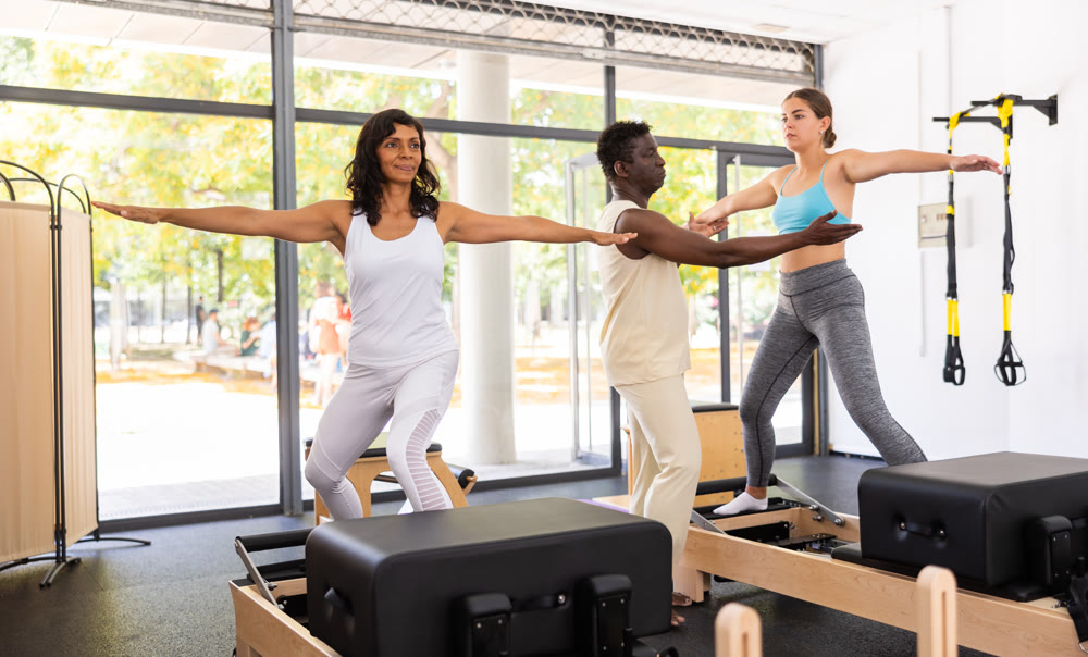 Attentive African American male trainer helping two positive women performing set of pilates