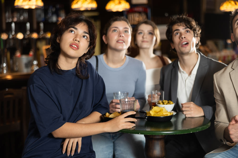 Group of young friends attentively watching match on TV and having fun together in beer pub