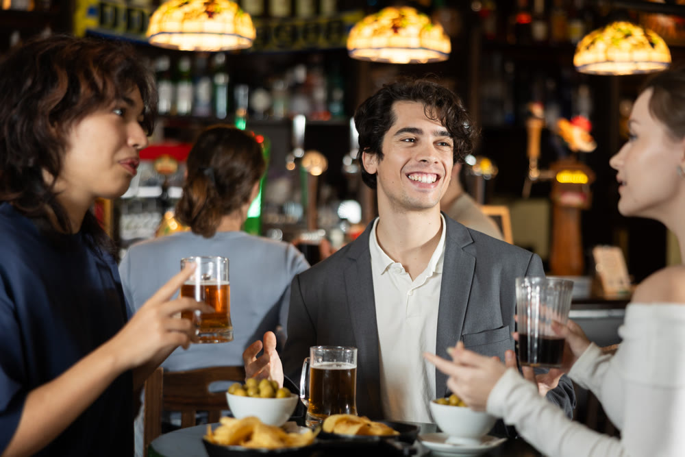 Positive cheerful young guy and two stylish girls enjoying cold beer in evening in pub
