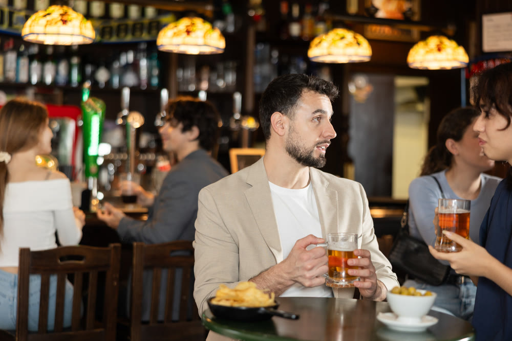 Couple of young guy and girl chatting, drinking beer and eating snacks at table in sports pub