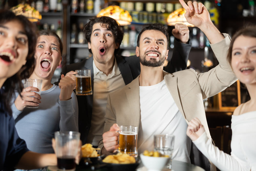 Fans watching match at a sports bar, cheering with drinks, energetic atmosphere, friendship and fun, people enjoying game together indoors
