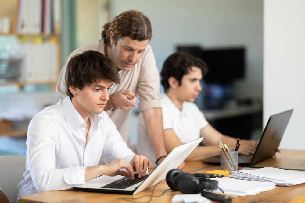 Several employees sit at desk during working day, working with laptop and paper documents.