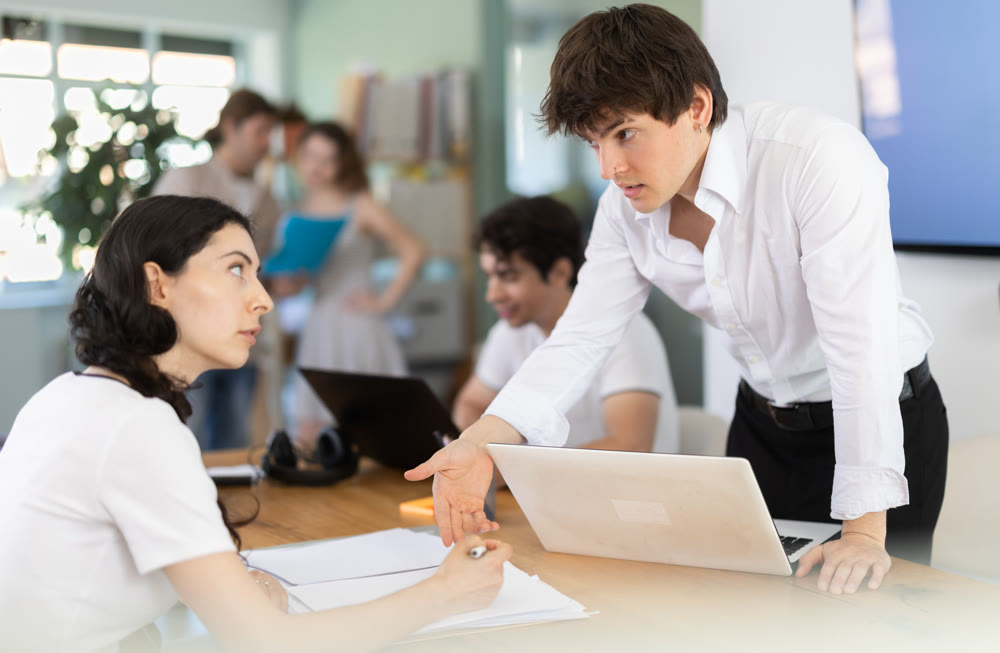 Several employees sit at desk during working day, working with laptop and paper documents.
