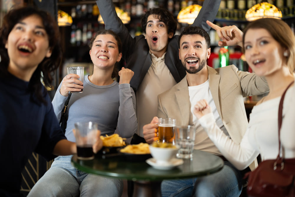 Fans watching match at a sports bar, cheering with drinks, energetic atmosphere, friendship and fun, people enjoying game together indoors
