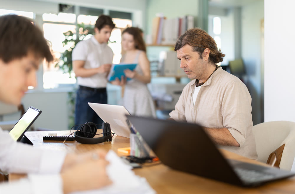 Several employees sit at desk during working day, working with laptop and paper documents.