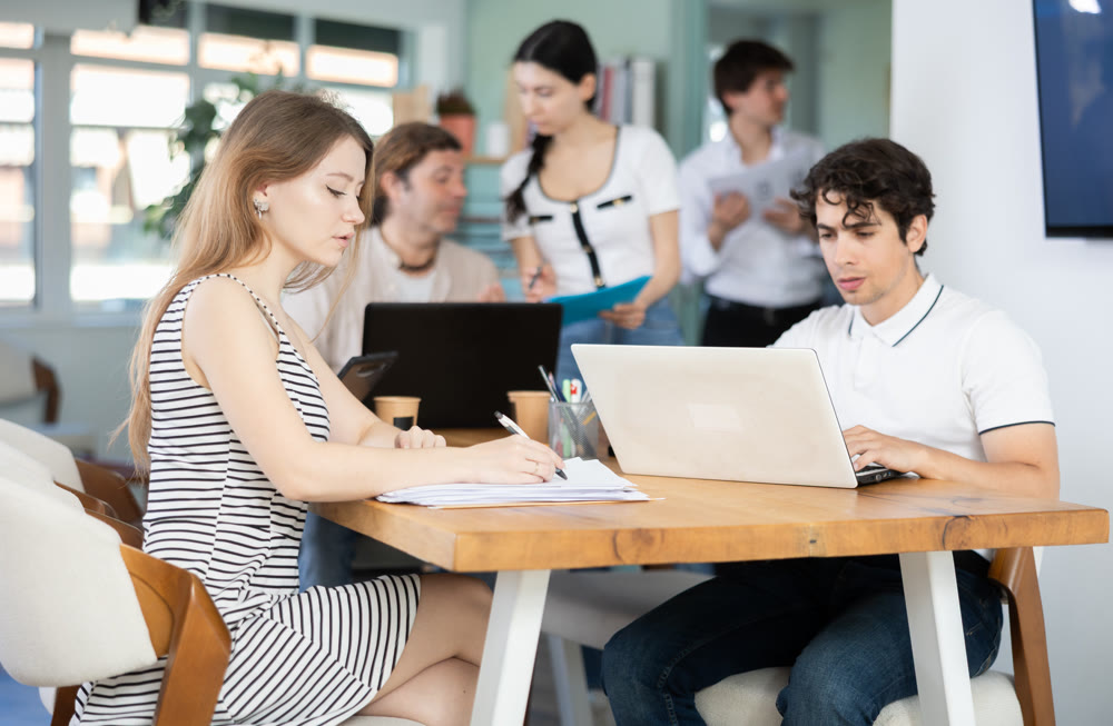 Several employees sit at desk during working day, working with laptop and paper documents.