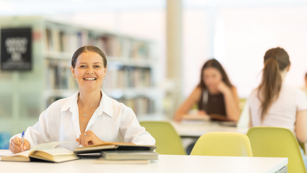 Female student sits at table in library and studies, reads books and takes notes
