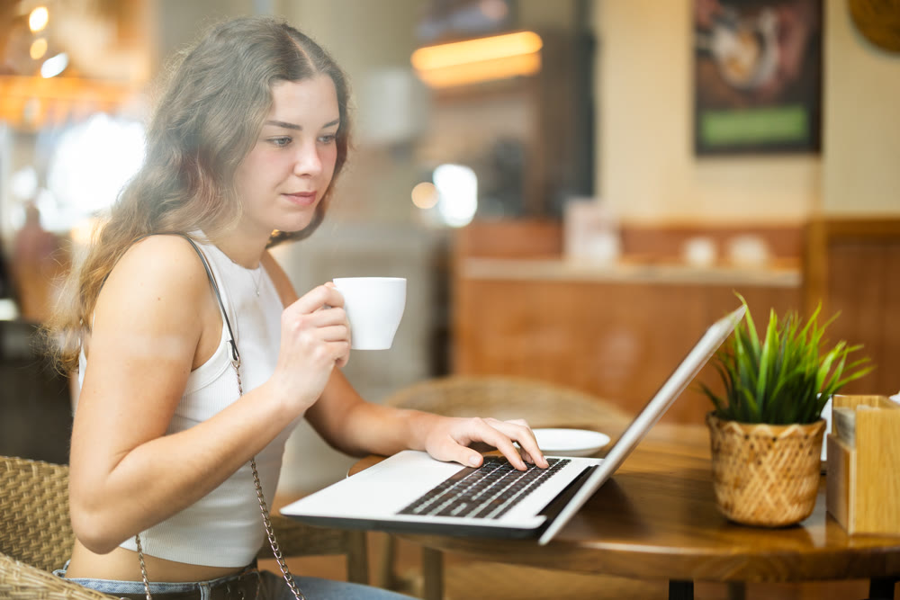 In cozy cafe hall, freelance girl works with laptop and drinks coffee.
