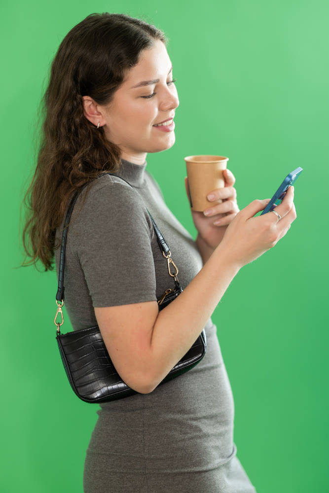 Young woman scrolling smartphone and drinking coffee in front of green screen