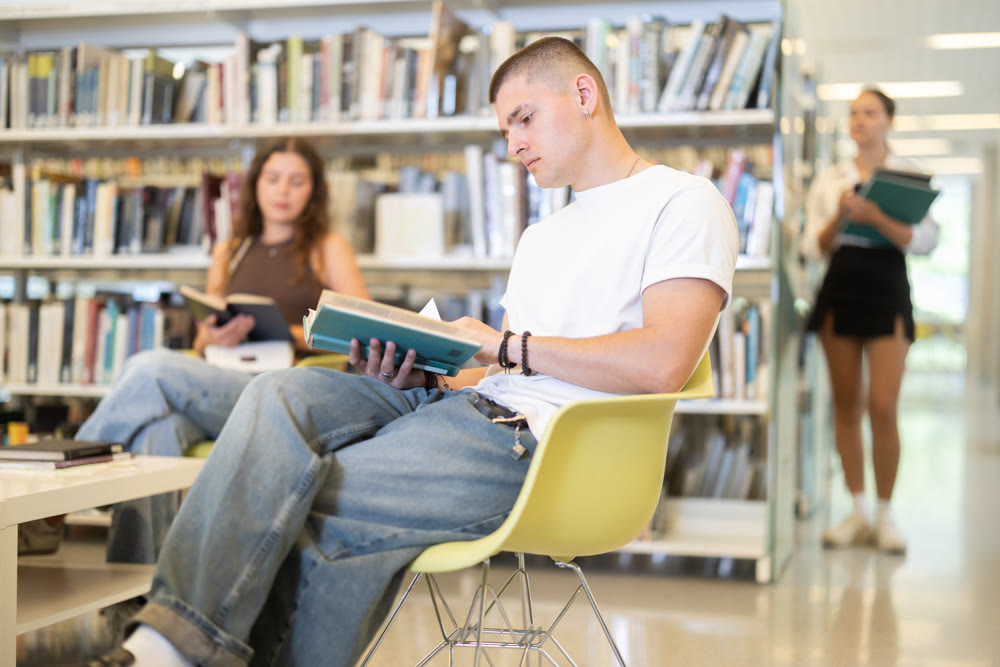 Male student reading book on chair in library