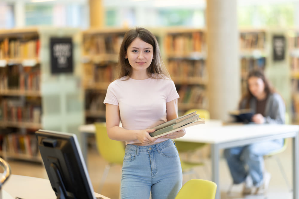 Portrait of positive student girl with books in her hands in university library