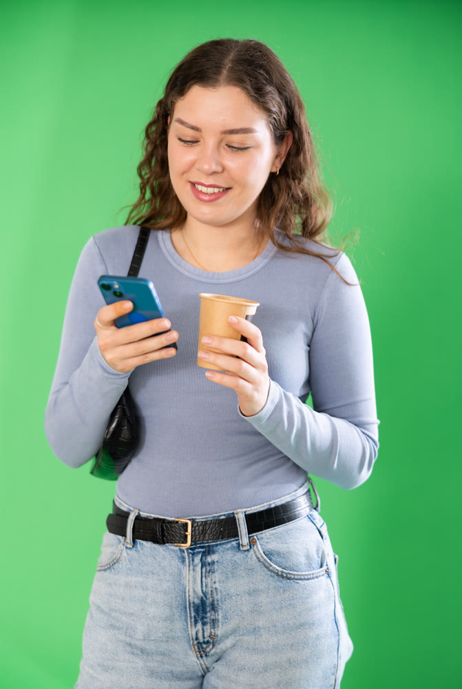 Young woman scrolling smartphone and drinking coffee in front of green screen