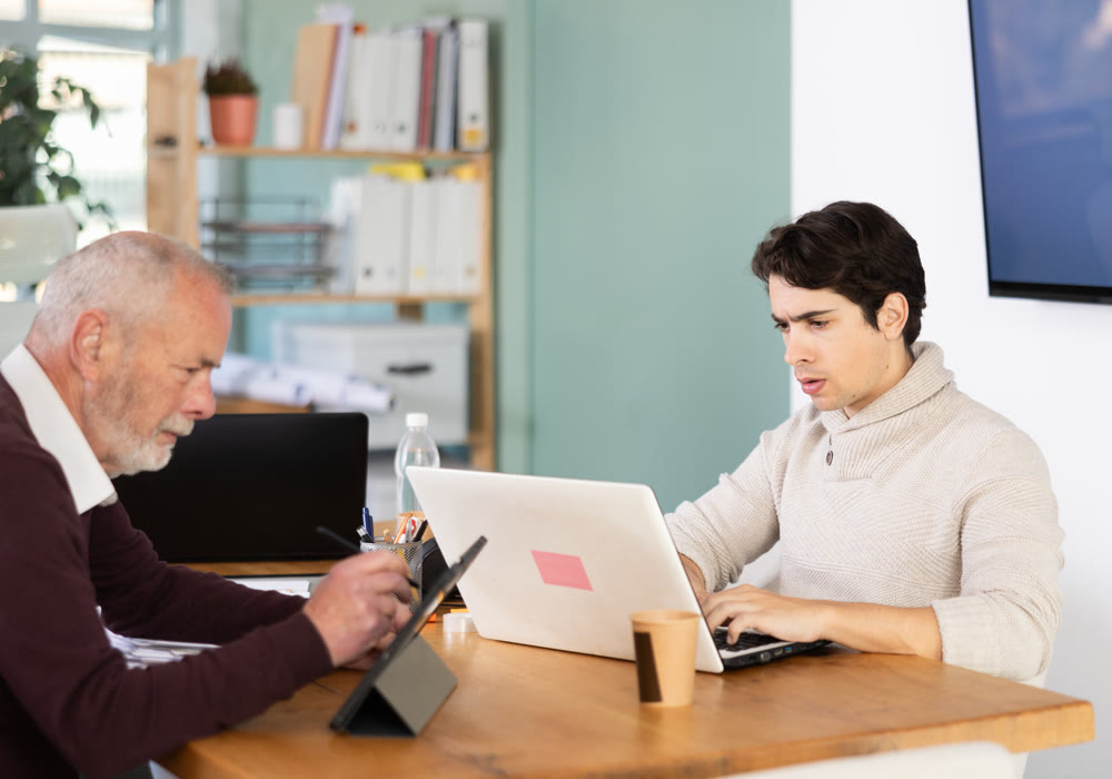 Elderly man working on tablet and young man on laptop in office