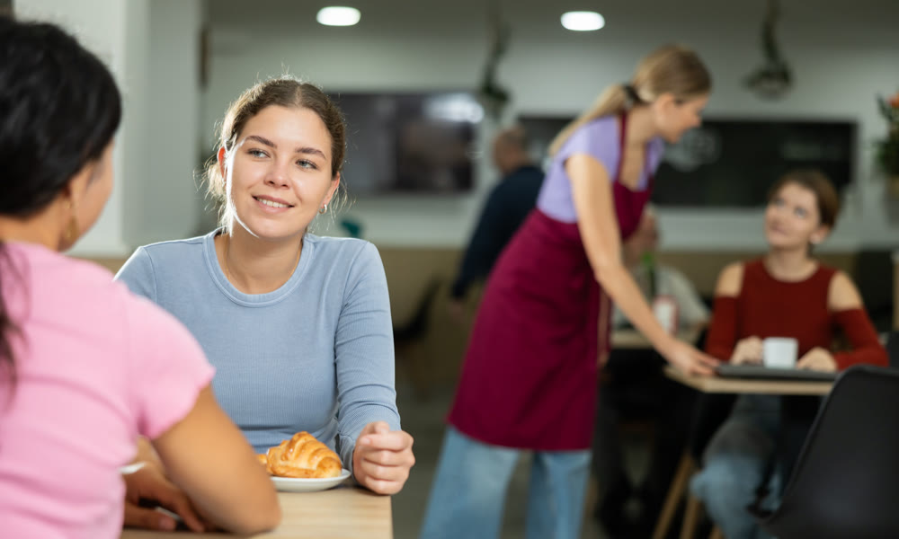 Young girl chatting with friend over coffee and croissant in cafeteria