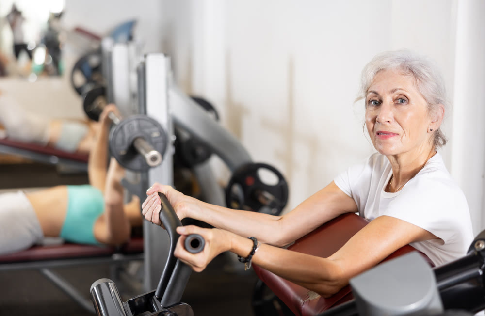 Slender senior woman work out muscles of arms and shoulders on simulator in gym of fitness club.