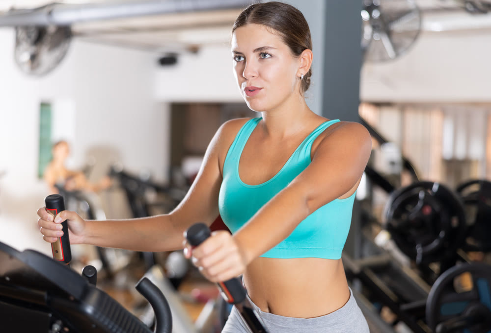 Slender athletic girl during cardio exercises on elliptical simulator in fitness center.