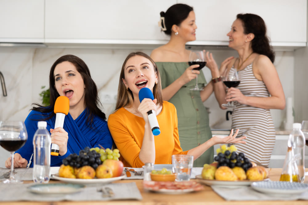 Group of female friends having fun at alcoholic bachelorette party in cozy living room, drinking red wine and singing together at home karaoke