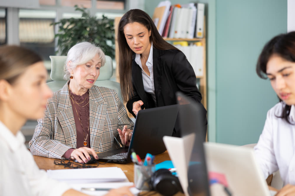 Young female leaned over to seated senior woman employee, helps with work and looks at laptop screen