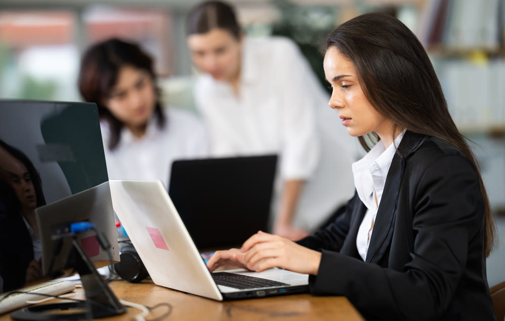 In office, young woman is working on laptop as part of group of colleagues.