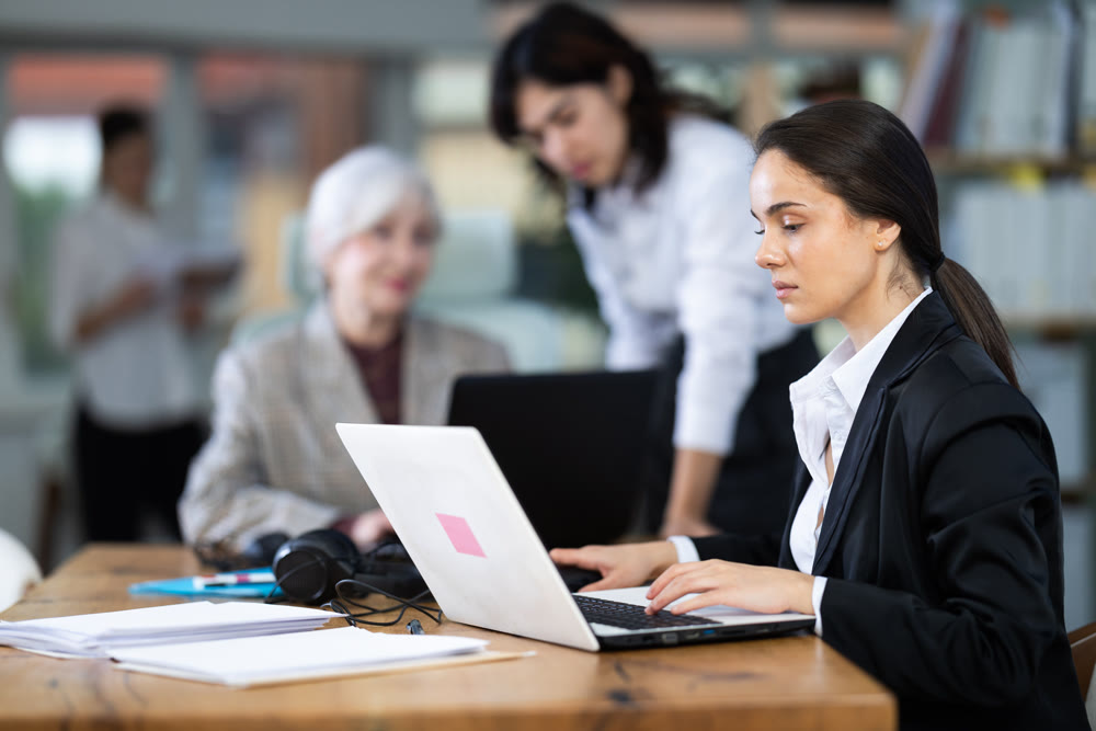 In office, young woman is working on laptop as part of group of colleagues.