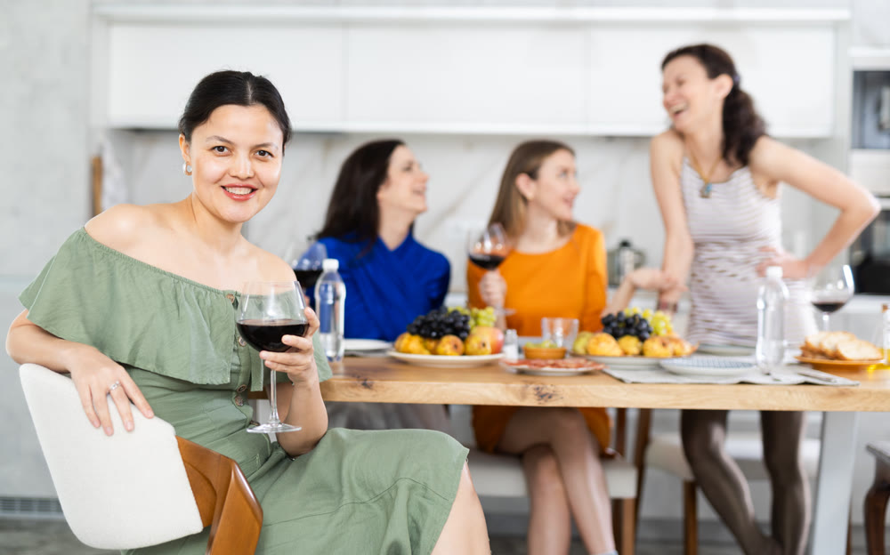 Portrait of joyful woman with glass of red wine against background of her friends at festive table