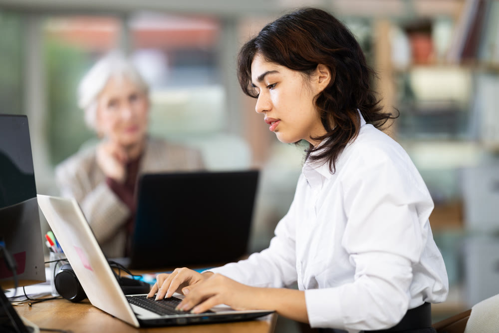 In office, young Asian woman is working on laptop as part of group of colleagues.
