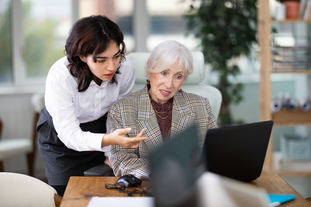 Asian girl leaned over to seated senior woman employee, helps with work and looks at laptop screen